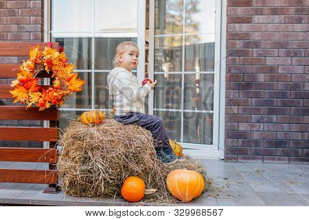 Adorable Smiling Caucasian Baby Toddler In White Knittes Jacket Sitting On The Haystack With Pumpkin