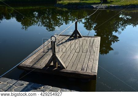 A Wooden Raft Is Tied To A Taut Rope. Self-service Ferry Across The River, Pond. People Get On And P