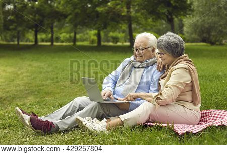 Happy Senior Couple Using A Laptop Computer While Sitting On A Park Lawn Together
