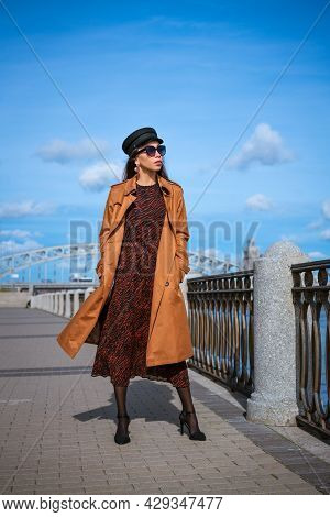 Beautiful Young Woman Of Caucasian Appearance In A Brown Raincoat And Dress Posing On The Embankment