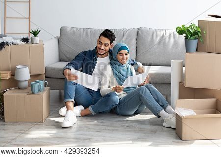 Cheerful Muslim Couple Sitting On Floor With Apartment Scheme