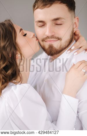 Groom With Her Lovely Bride Hug Each Other And Kiss In Cheek. Happy Wedding Couple In Front Of Grey 