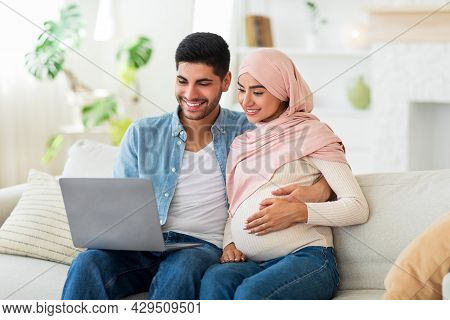 Loving Pregnant Muslim Couple Choosing Furniture For Baby Room Online, Using Laptop On Sofa In Livin