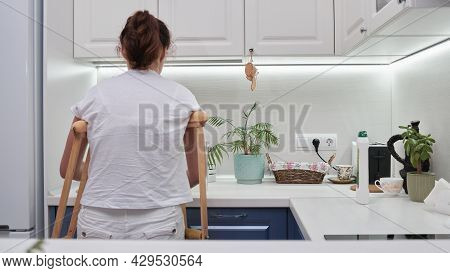 Woman On Crutches Washes The Dishes In The Kitchen. Temporary Limitation Of The Physical Capabilitie