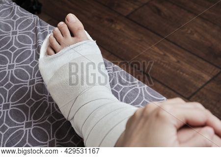 Woman Sits On The Bed And Holds A Broken Leg. The Leg Is In A Plaster Cast. Home Rehabilitation.