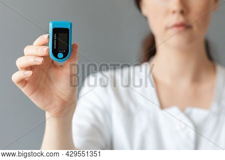 Blurred Portrait Of Doctor Shows A Pulse Oximeter. Gray Background. The Concept Of Health Care And C