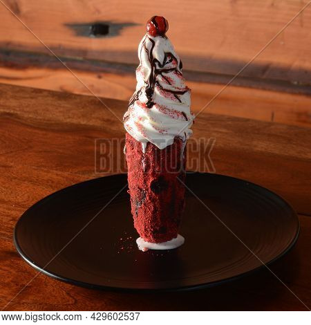 Red Velvet Milkshake Topped With Ice Cream And Cherry Served In A Plate Over A Rustic Wooden Backgro