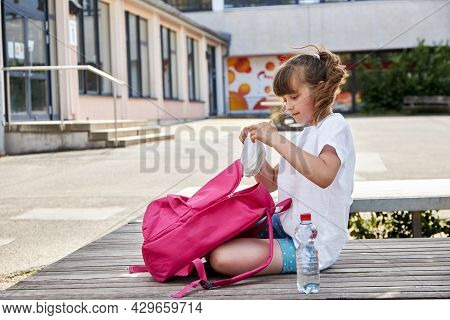 A Schoolgirl Gets Out Of Her Backpack Her Lunch, A Sandwich In The Schoolyard. Children Eat At Schoo