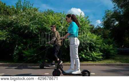 Young Happy Couple Driving Electric Scooters In City Park At Sunny Summer Day. First Date