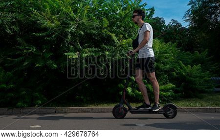 Young Man Dressed In White T-shirt Riding Black Electric Kick Scooter In Green Cozy Park..