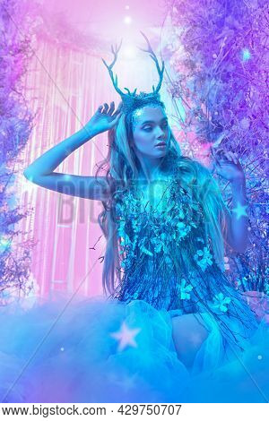 Winter fairy tale. Beautiful horned wood fairy in a chic dress in a magical forest. Fantasy world in light blue and light purple lighting. 