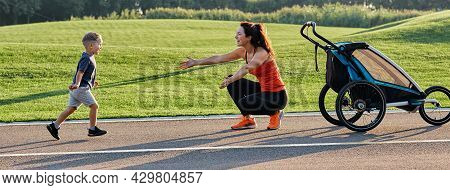 Active Mother With Child Using Jogging Stroller For Running At Park. Happy Mother And Her Son Having