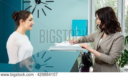 Lady Office Manager In White Blouse Shows Registration Book To Woman Client Standing At Reception In
