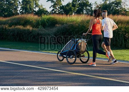Running Young Family With Baby Jogging Stroller In Motion Enjoying Workout And Summer Day In City Pa