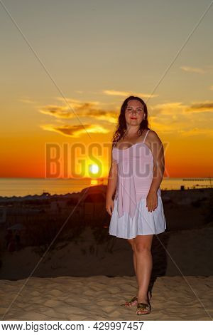 Woman In A White Dress On The Beach Against The Background Of The Sea At Sunset.