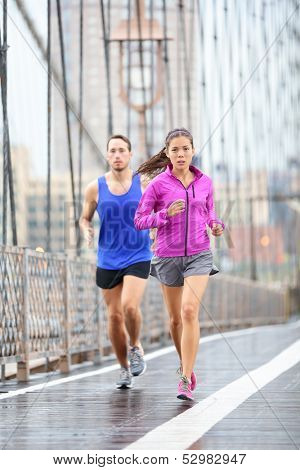 Running couple. Runners jogging outside in rain. Woman and man runner athletes training outdoor for marathon on Brooklyn Bridge, New York City, USA. Asian female and Caucasian male fitness sport model