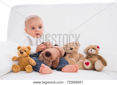 Amongst toys: cute baby sitting on white sofa with teddy bears 