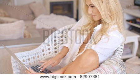 Pretty sexy blond woman relaxing in a white wicker chair in her living room working on a laptop computer with a smile  side view