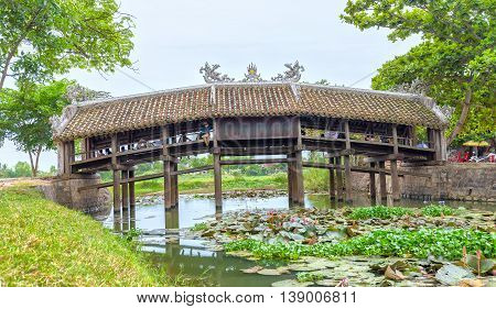 Thua Thien Hue, Vietnam - June 21st, 2015: Architecture bridge with wooden tile roofing structures on like a house on river to attract tourists to relax, below lilies blooming creating beauty idyllic countryside in Thua Thien Hue, Vietnam