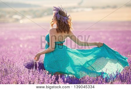 Happy pregnant brunette woman with long curly hair,large belly,dressed in a long dress turquoise,a hand holding a basket of flowers ,on her head is a wreath of field of lavender,picking flowers on a summer meadow mountain lavender