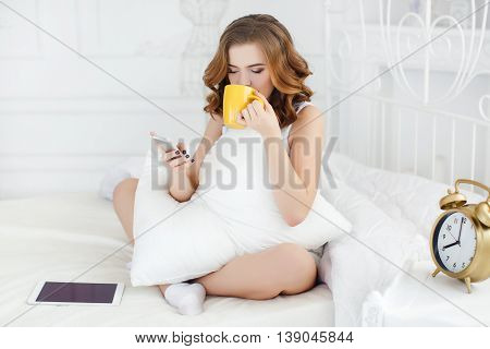 Young beautiful woman with wavy red hair and gray eyes,light makeup and cute smile,dressed in a white shirt,sits in the morning on a white bed in the bedroom,reading a message on a white smartphone in hand holds a yellow Cup with strong coffee