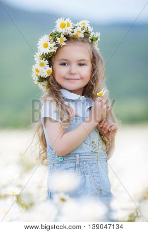 Beautiful little girl with long curly blond hair,cute smile,in a light blue denim overalls,a white wreath of fresh flowers,holding hands in a flower field daisies,enjoying nature in a flowering meadow white in summer