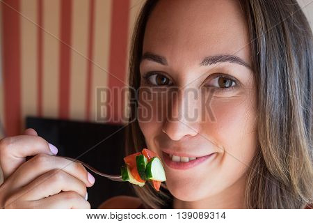 The girl is holding a fork at the mouth with slices of tomatoes and cucumbers the girl stands in the kitchen looking at the camera and smiling taste of summer salad