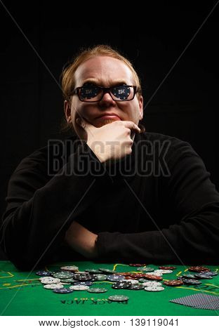 Beautiful young red-haired man in glasses and shirt with long sleeves sitting at poker table with cards and chips on it inserting two chips before his eyes at black background.