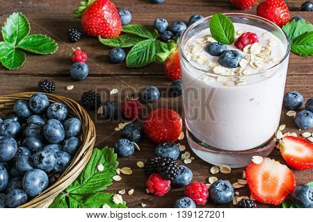 glass of yogurt with whole fresh blueberries blackberries raspberries and oatmeal on old rustic wooden table with wicker bowl and many ripe berries. Closeup Detail.
