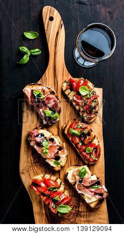 Brushetta set for wine. Variety of small sandwiches with prosciutto, tomatoes, parmesan cheese, fresh basil and balsamic creme served with glass of red wine on rustic wooden board over dark background, top view