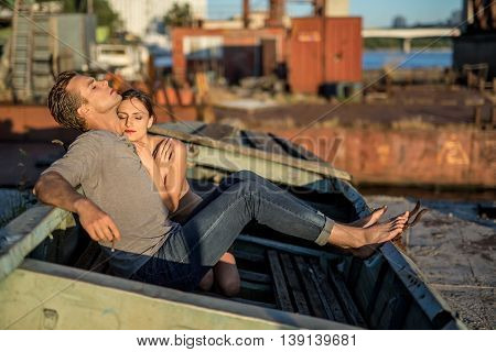Attractive couple with closed eyes in the old boat outdoors in the industrial zone. Barefoot guy in a gray T-shirt and blue jeans smokes a cigarette. Nude girl cuddled up to him. Horizontal.