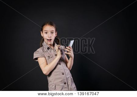 Portrait of surprised excited girl dial on touch screen of mobile phone at black background. Female child using internet in cell phone. New generation devices, communication