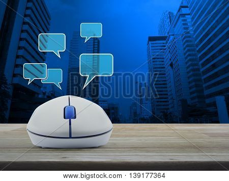 Wireless computer mouse with social chat sign and speech bubbles on wooden table in front of city tower background Social network concept