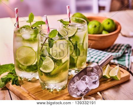 Alcohol cocktail. On wooden boards three glasses with alcohol cocktail and ice cubes. Drink number hundred and ninety two cocktail mohito with straw and lime and mint leaf. Country life. Indoor.