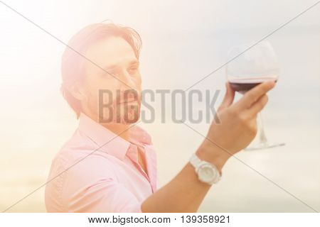 Toned picture of sommelier analyzing glass of red wine isolated on blue sky background. Black-haired mature man happy looking at glass.