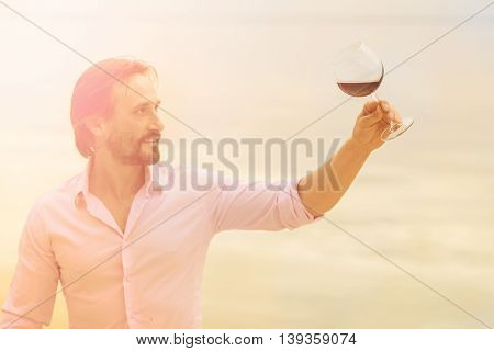 Toned picture of satisfied mature professional sommelier looking at glass with red wine. Handsome man in pink shirt drawing aside glass.