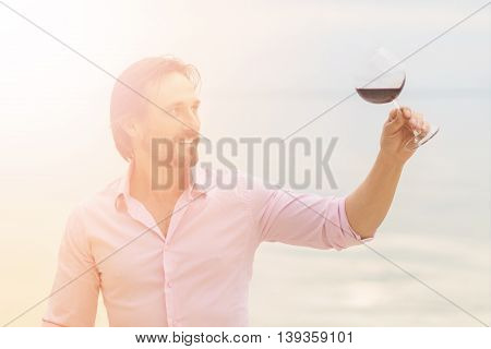 Confident sommelier. Toned image of thoughtful young sommelier holding glass of red wine and looking at it while staying near by sea.