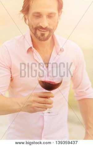 Confident and happy sommelier. Toned image of bearded mature man in pink shirt tasting red wine and looking at glass isolated on sea.