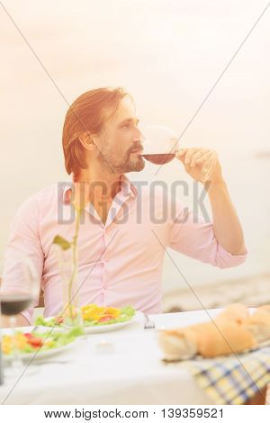 Toned portrait of handsome man drinking red wine while resting in restaurant or cafe by sea. Bearded man in pink shirt spending time abroad or outskirts.