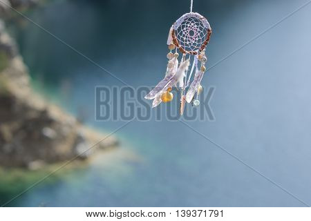 Handmade pink native american dream catcher on background of rocks and lake. Tribal elements feathers shells lace