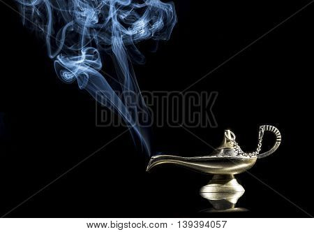 Magic lamp on black background from the story of Aladdin with Genie appearing in blue smoke concept for wishing, luck and magic.