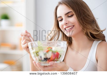 So tasty. Cheerful delighted beautiful woman holding bowl and eating salad while resting at home