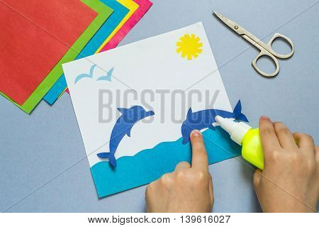 Child glues a dolphin out of paper making the applique on the marine theme. The idea for children's creativity an art project made of paper. Sheets of colored paper glue scissors