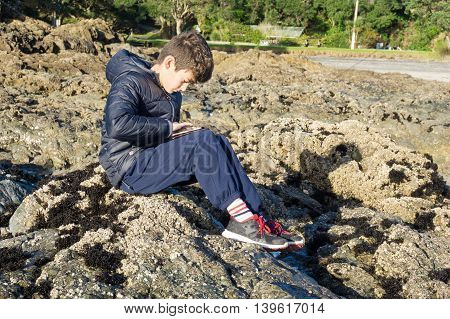 Boy on rocks playing on mobile device Waipu Cove