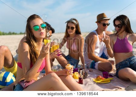 Positive emotions. Cheerful attractive woman sitting on the blanket and blowing soap bubbles while having a picnic with her friends