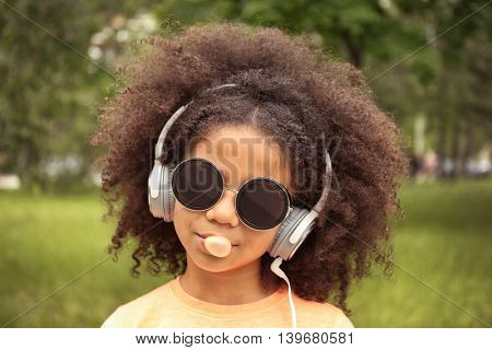 Afro-American little girl with sunglasses and headphones chewing bubblegum in park
