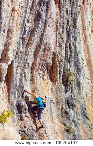 Blond Female Climber ascending Vertical Orange Rocky Wall unusual shape of Mountain Range on background Outdoor Sunshine