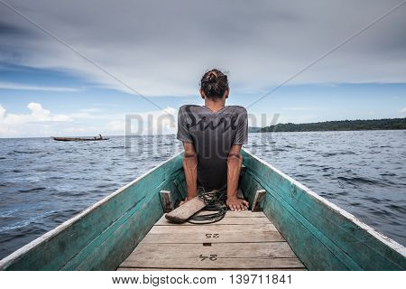 Photo of Man Sitting Natural Wood Long Tail Boat Caribbean Ocean. Clear water and blue sky with clouds. Horizontal