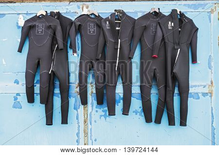 Wetsuits in children's sizes dry in the sun on a door of blue which is rusting in places due to salt corrosion of sea air.