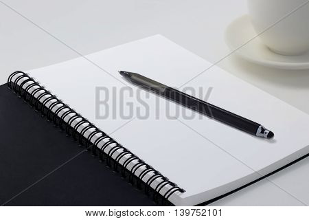 Blank notebook on white desk background with a pen on top and coffee cup on the right side.